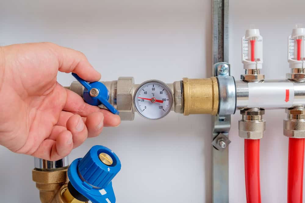 A hand is adjusting a blue valve on a heating system, part of routine boiler maintenance, with a pressure gauge displaying the current pressure. The system includes red pipes and other components such as additional gauges and connectors, indicating a complex plumbing or heating setup.