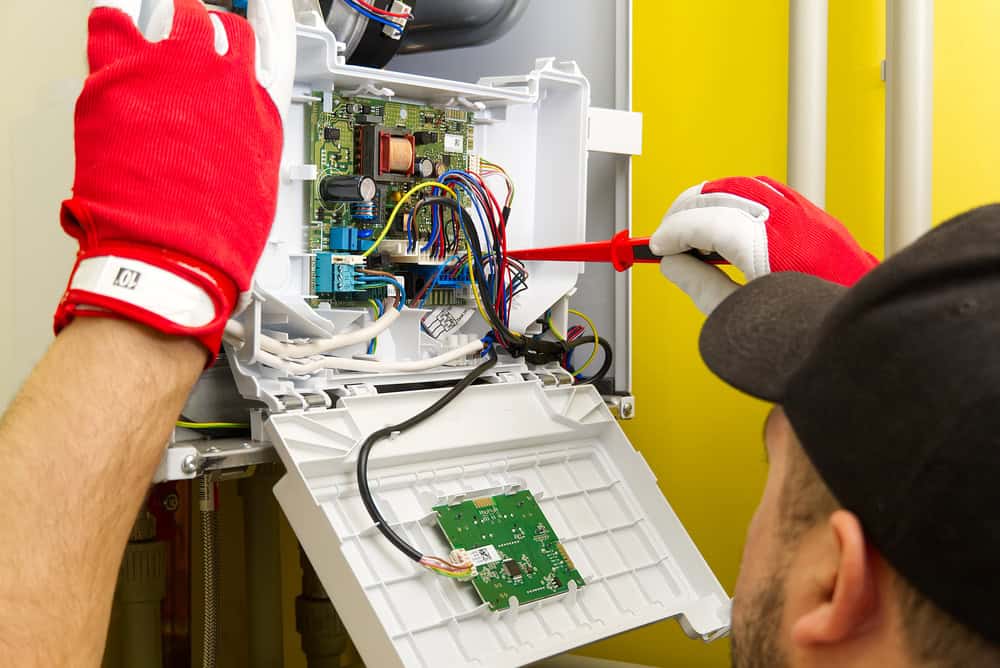 A technician wearing red gloves is working on an open electrical panel, using a screwdriver to adjust wires and components. The panel, related to HVAC services, is mounted on a bright yellow wall, showcasing various colorful wires and circuit boards.