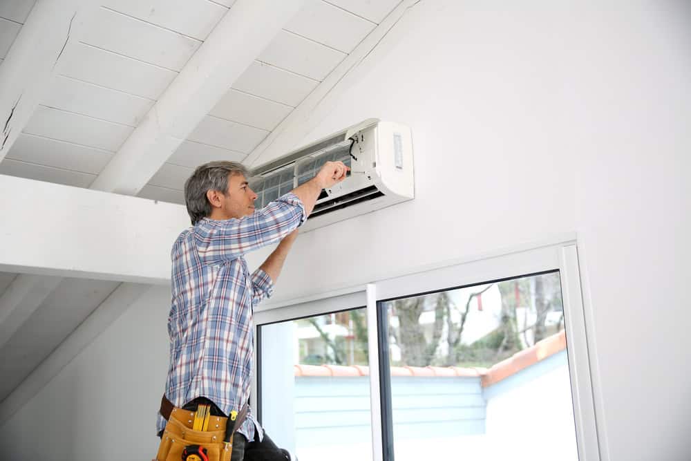 A person wearing a plaid shirt is standing on a ladder, performing HVAC services by repairing or installing a wall-mounted air conditioning unit inside a room with a slanted ceiling and large windows. Tools are visible in a tool belt around their waist.