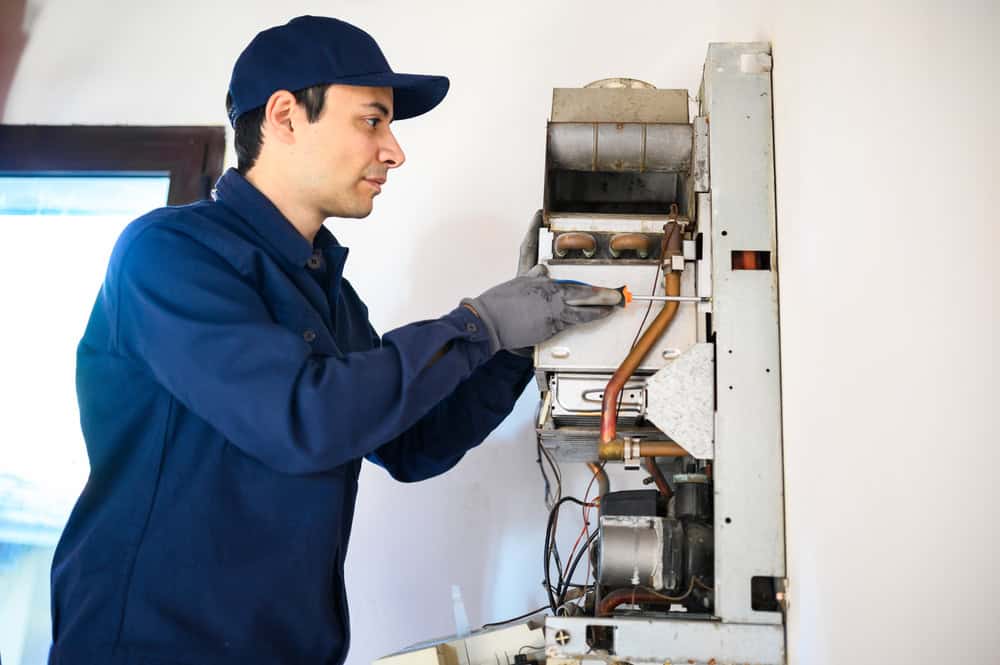 A technician in a blue uniform and cap repairs a wall-mounted heating unit, showcasing his expertise in boiler maintenance. He uses hand tools and wears gray gloves for protection. The unit's inner components, including wires and pipes, are exposed. The background features a white wall and a window.