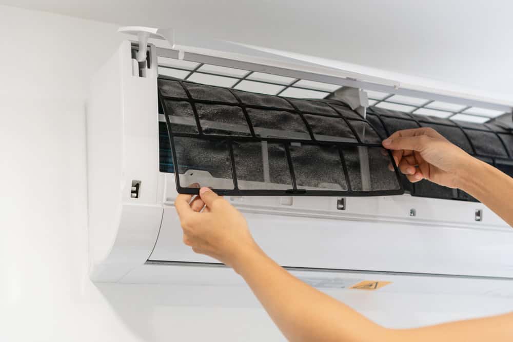 A person is engaged in AC repair, cleaning the filter of a split air conditioning unit mounted on a wall. The individual's hands are holding and removing the dust-filled filter from the open AC unit. The filter appears to be dirty and in need of cleaning.