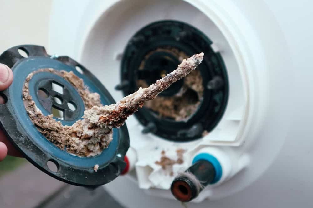 Close-up of a heavily corroded water heater element being removed from a tank. The element is encrusted with scale and mineral deposits, and the tank’s opening shows signs of rust and buildup. Red and blue inlet/outlet ports are visible on the heater, highlighting the importance of regular boiler maintenance.