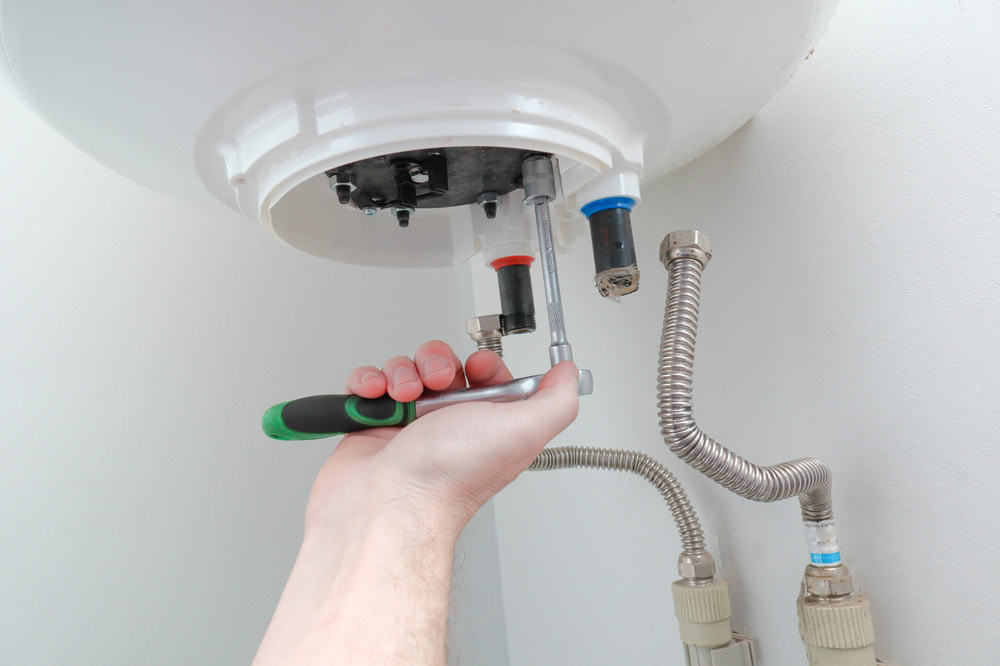 A hand using a wrench to adjust connections on the underside of a white water heater, demonstrating essential HVAC Services. Two flexible metal hoses are attached to the heater, and the hand holds the wrench near the blue and red water supply lines. The surroundings are clean and white.