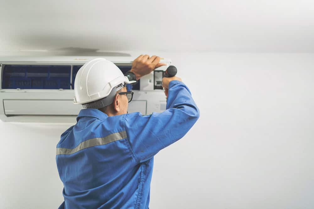 A technician wearing a white hard hat and blue work uniform is performing AC repair on a wall-mounted unit. He is using a screwdriver to fix the unit's interior components. The background is a plain white wall, reflecting the focus on meticulous HVAC services.