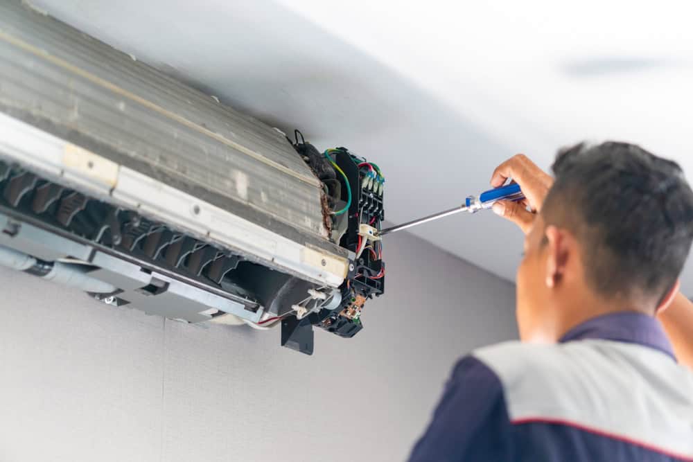 A person is using a screwdriver to repair an air conditioning unit mounted on a wall. Focused on the internal components of the open unit, various wires and parts are visible. The scene, reminiscent of intricate AC repair tasks, unfolds against a plain, light-colored wall.