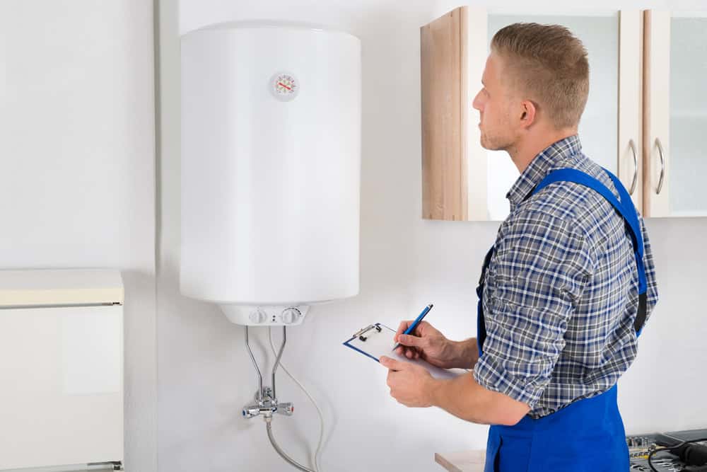 A man in blue overalls and a checkered shirt is inspecting a wall-mounted water heater, possibly conducting boiler maintenance. He holds a clipboard and pen, taking notes. The setting appears to be a residential kitchen with white walls and cabinets.