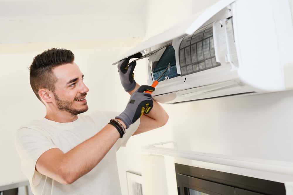 A smiling person is wearing gloves and using tools to repair or maintain a wall-mounted air conditioning unit. The individual, dressed in a white t-shirt and standing indoors, has a focused look on their AC repair task.
