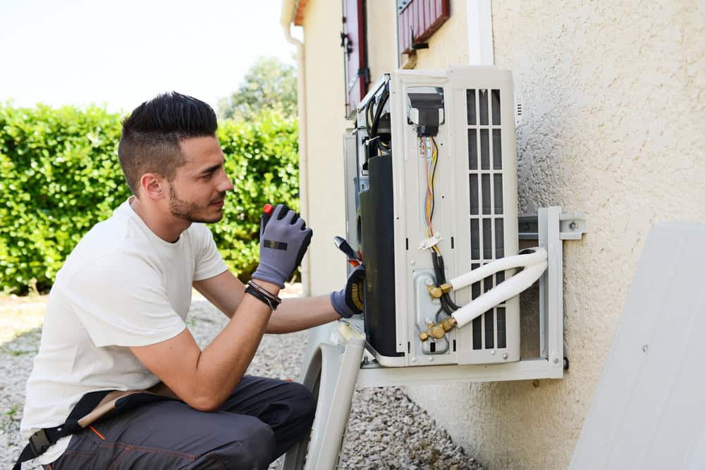A technician wearing a white shirt and gray gloves is working on an outdoor air conditioning unit mounted on the exterior wall of a building. He holds a tool in his right hand, maintaining focus on the AC repair. In the background, there are bushes and trees.