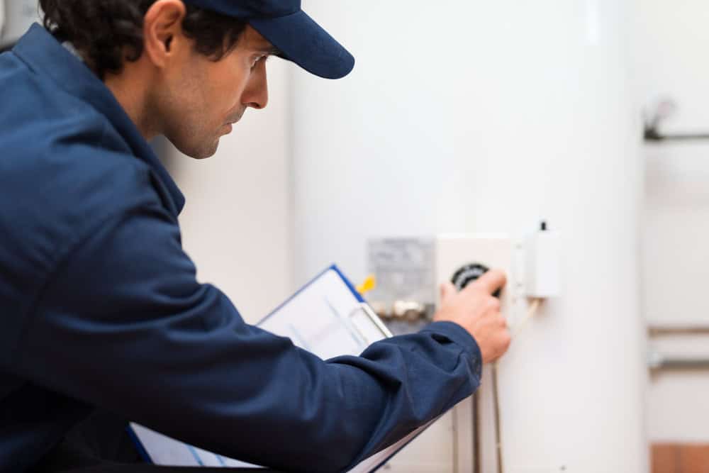 A person wearing a blue uniform and cap is inspecting a white appliance, likely a heater or boiler, while holding a clipboard with paperwork. They appear to be adjusting a dial or control as part of routine boiler maintenance.