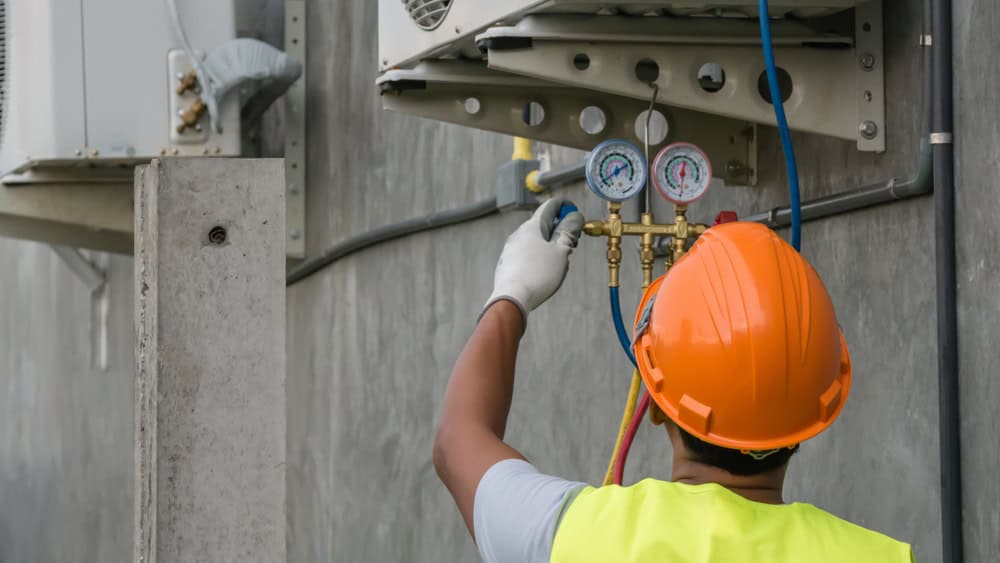 A worker wearing a reflective vest, white gloves, and an orange hard hat is adjusting gauges on an air conditioning unit mounted on a gray concrete wall, showcasing professional HVAC services. The worker is viewed from behind.