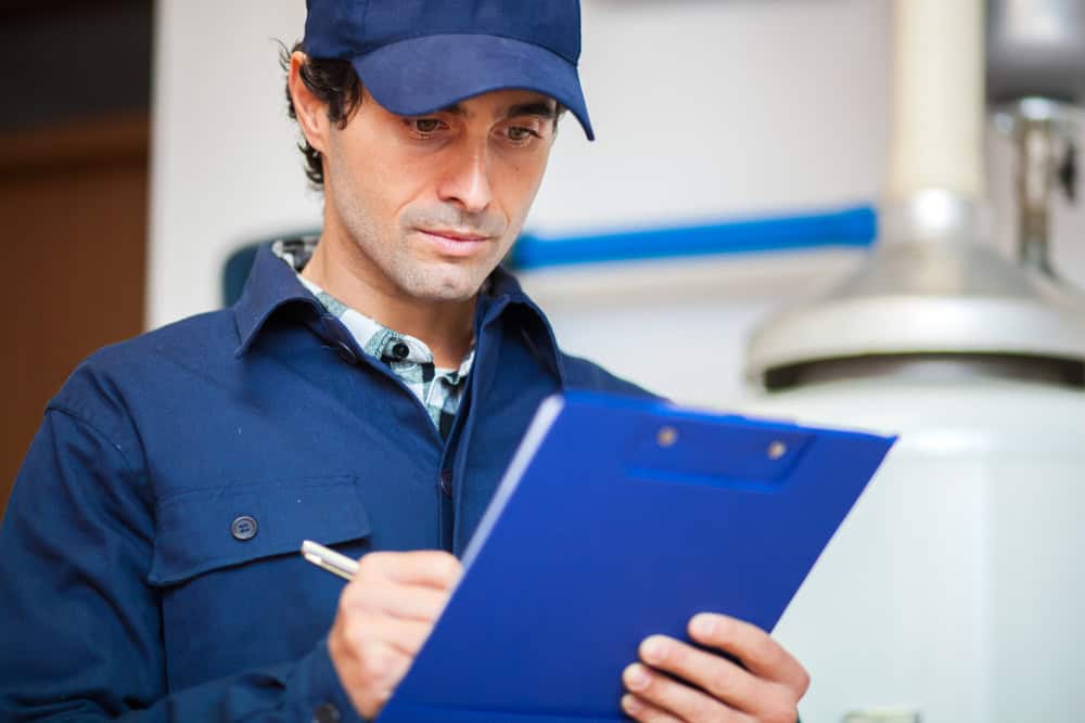 A man in a blue uniform and cap is holding a blue clipboard and writing with a pen. He is focused, seemingly conducting an HVAC services inspection in an industrial or work setting.