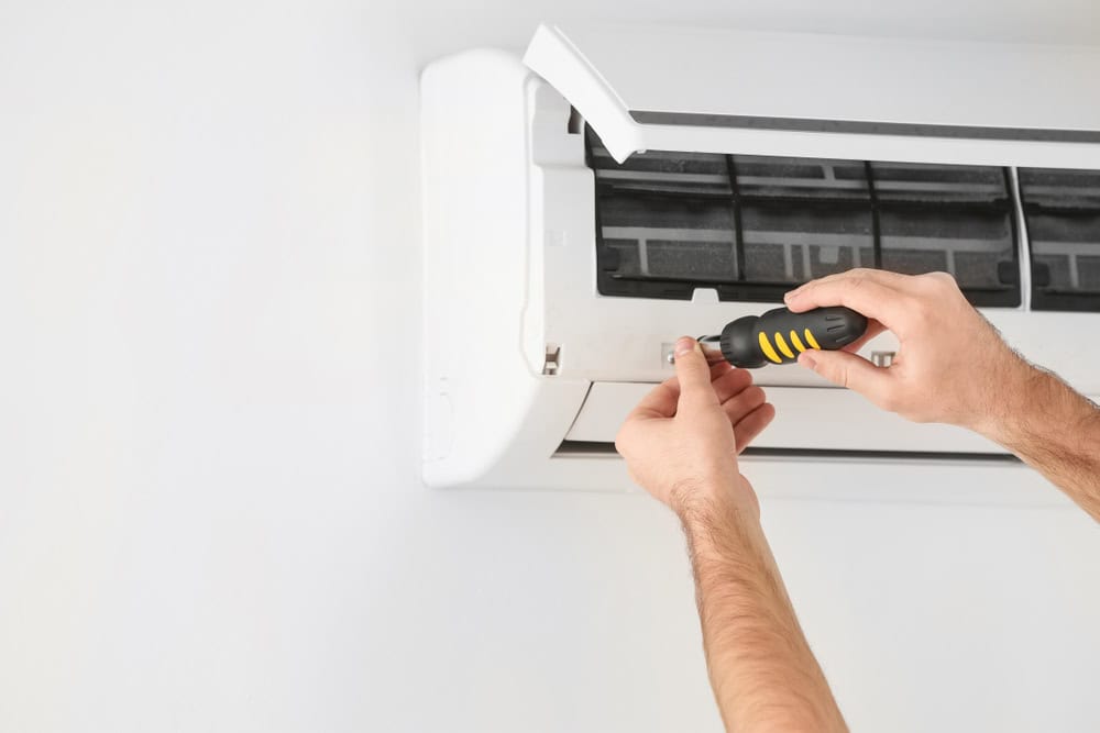 A person using a screwdriver to repair or install a wall-mounted air conditioning unit. The person's hands and the screwdriver are visible, with the front panel of the air conditioning unit partially opened. This meticulous task highlights the essential role of HVAC Services in maintaining home comfort.