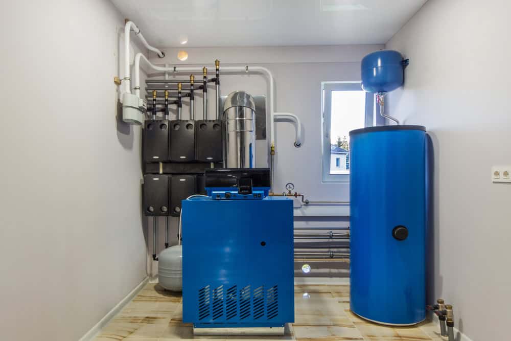 A utility room with a blue water heater and boiler system, surrounded by various pipes and valves, designed for streamlined boiler maintenance. The room has light-colored walls and a small window letting in natural light. The floor is tiled, and several black control units are mounted on the wall.