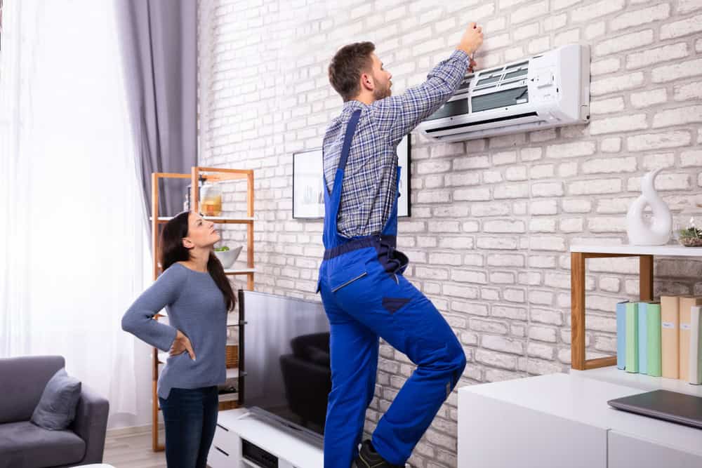 A technician in blue overalls stands on a small ladder, engaged in AC repair on an air conditioner mounted on a brick wall. A woman watches him with her hands on her hips in a modern living room featuring a couch, shelves, and various decor items.
