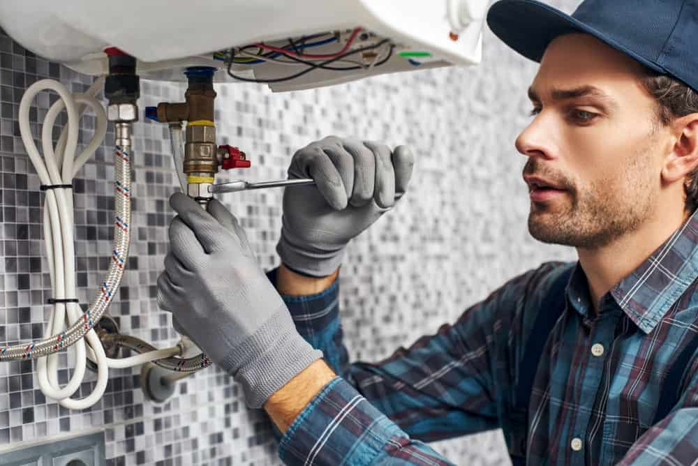 A man wearing gloves and a cap is diligently repairing a pipe in a tiled room, using a wrench to tighten a fitting. Wires and other plumbing components surround him as he works, showcasing his expertise in HVAC services.