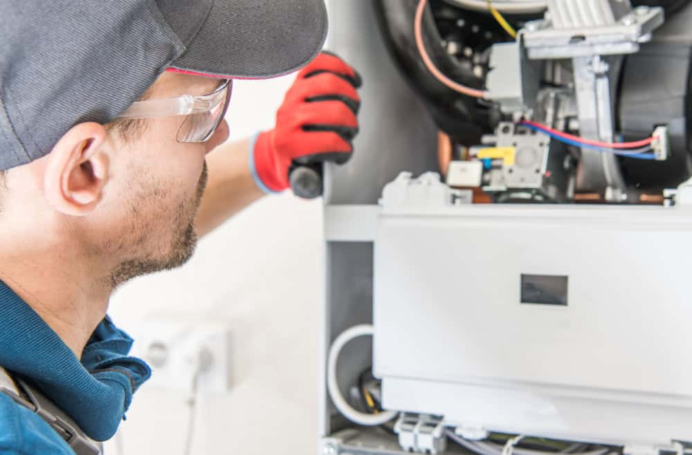 A maintenance technician wearing safety glasses, a cap, and a red glove is inspecting or repairing the inside of a household appliance or electrical panel. The technician, possibly conducting boiler repair, is looking intently at the components, with tools and wires visible in the panel.