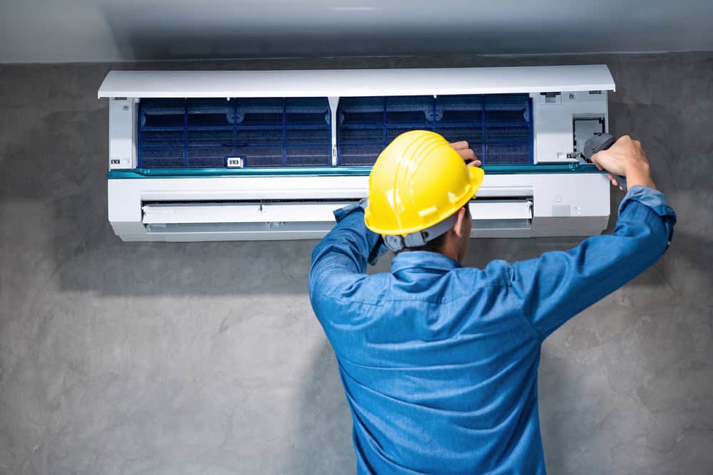 A person wearing a blue shirt and a yellow hard hat works on an open wall-mounted air conditioning unit. They are using a screwdriver to adjust or fix the device against a gray wall, showcasing their HVAC services expertise.