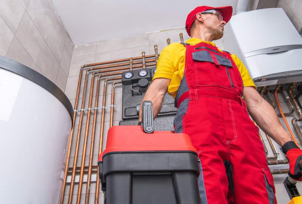 A technician in a red and yellow uniform and cap holds a toolbox while standing in a boiler room. Behind him, there are various pipes and heating equipment, including a large cylindrical water heater and a wall-mounted boiler. The room has tiled walls, indicating ongoing HVAC services or potential boiler repair.