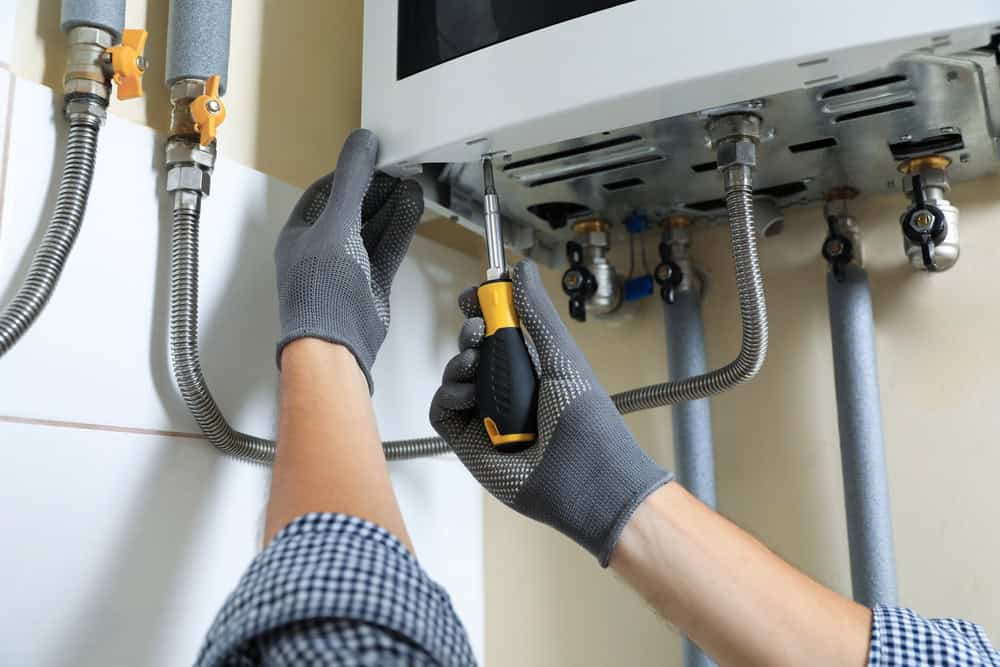 A close-up of a person in gloves using a screwdriver to repair or install a gas boiler. Several pipes and connections are visible around the boiler unit, highlighting the attention to detail involved in boiler maintenance. The person is wearing a checkered shirt.