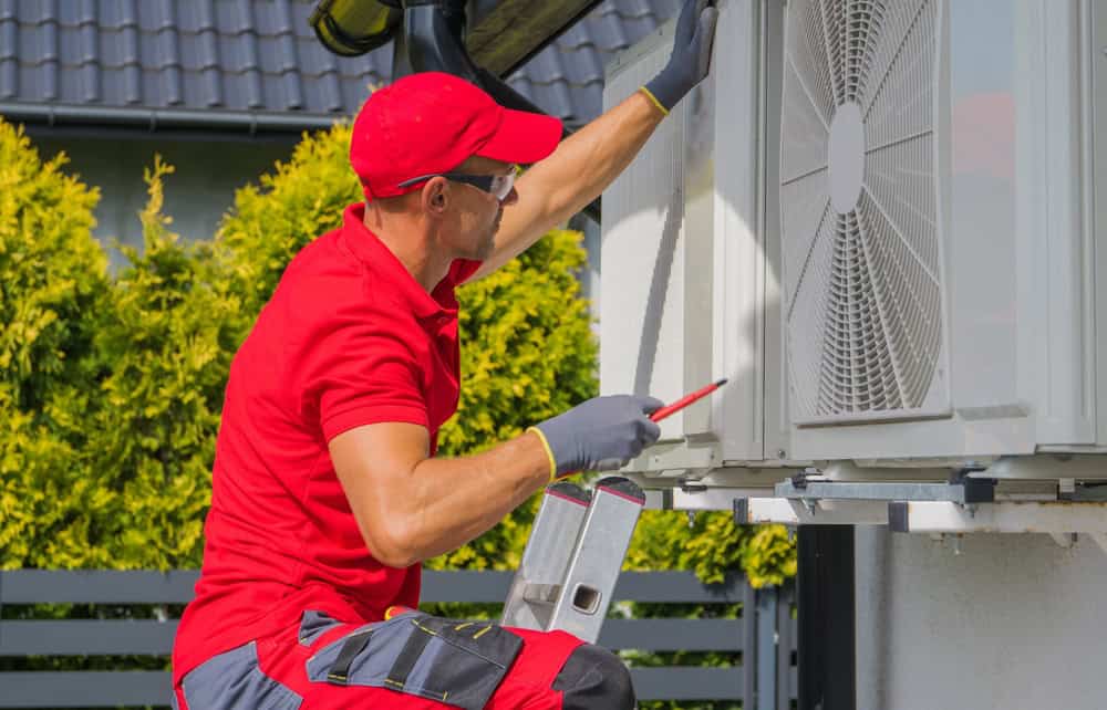 A technician in a red uniform and cap, wearing gloves and safety glasses, installs or repairs an outdoor air conditioning unit mounted on a house wall. The technician, focused on AC repair, is holding a tool and standing on a small step ladder. Shrubs and a fence are in the background.