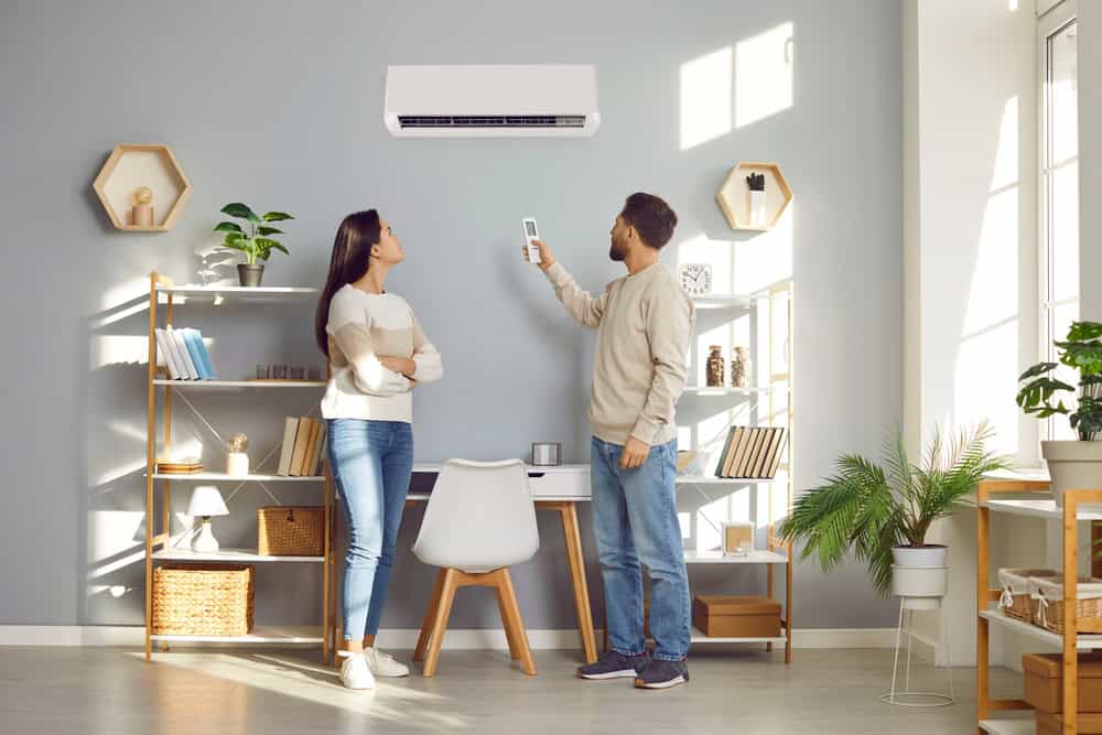 A man and woman stand in a modern, neatly organized living room with a grey wall. The man is pointing a remote control towards an air conditioning unit after its recent AC repair. The room features a bookshelf, desk, and various plants. Natural light fills the space.