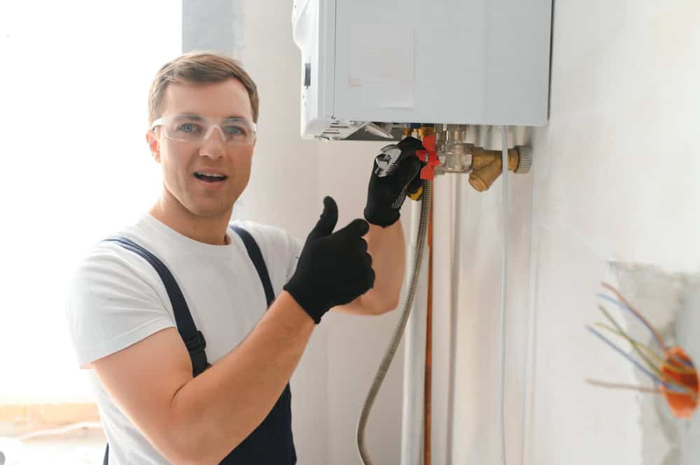 A man wearing safety glasses, a white t-shirt, black gloves, and overalls is working on a wall-mounted device, possibly during boiler installation. He is giving a thumbs-up with one hand and holding a tool in the other.