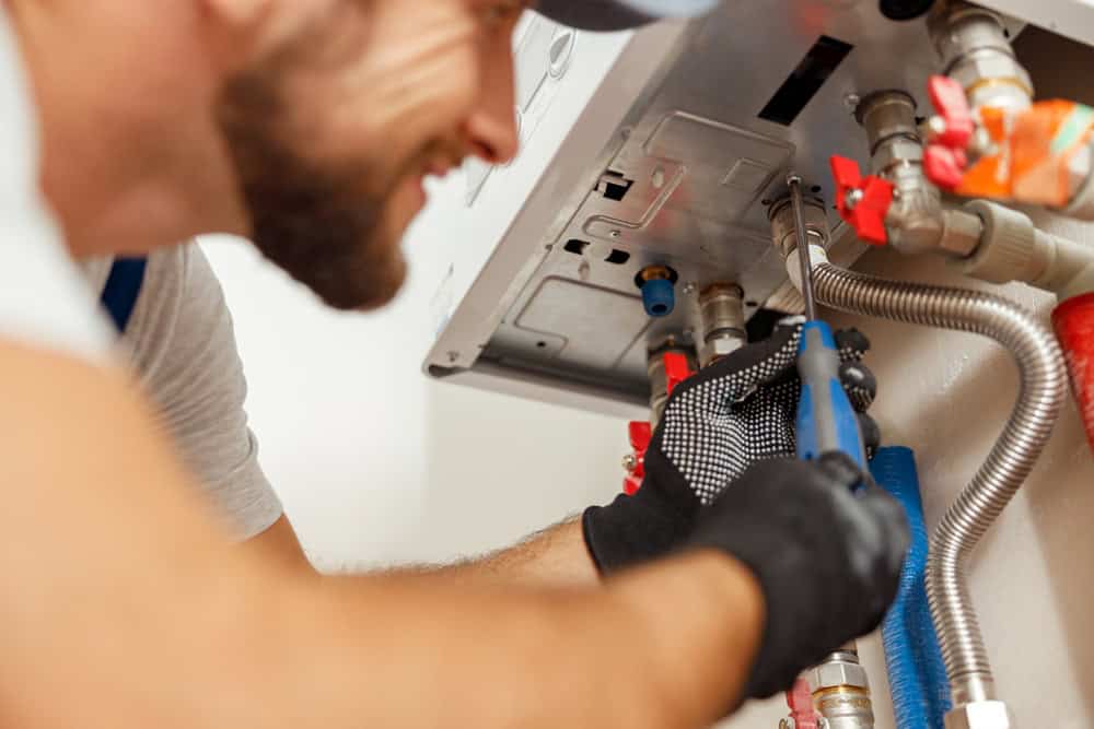 A plumber wearing a gray cap and black gloves uses a wrench to work on pipes and connections underneath a large, mounted appliance. The scene is up close, focusing on the plumber's hands, tool, and the mechanical components, illustrating meticulous boiler maintenance in progress.