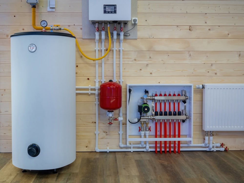 A boiler room setup showcasing a cylindrical water heater, connected pipes, a red expansion tank, and a wall-mounted control panel. Various pipes and valves are neatly arranged against a wooden wall, with a segment of hardwood floor in view—a perfect example of efficient Boiler Maintenance.