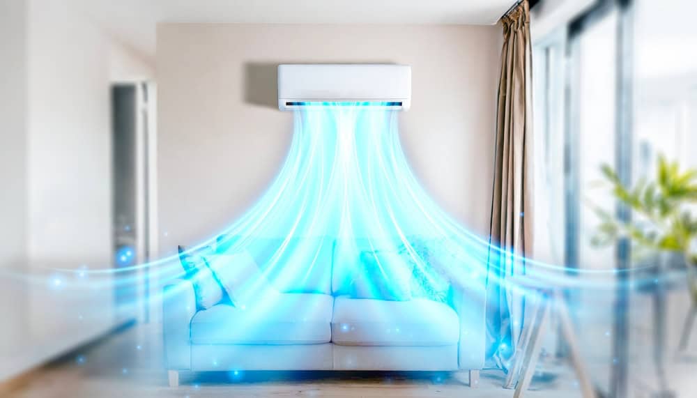 An air conditioning unit, recently serviced for AC repair, is mounted on the wall of a modern living room, releasing cool air depicted as blue streams. A white sofa sits directly below, and large windows with curtains line the right side of the room, allowing natural light to flood in.