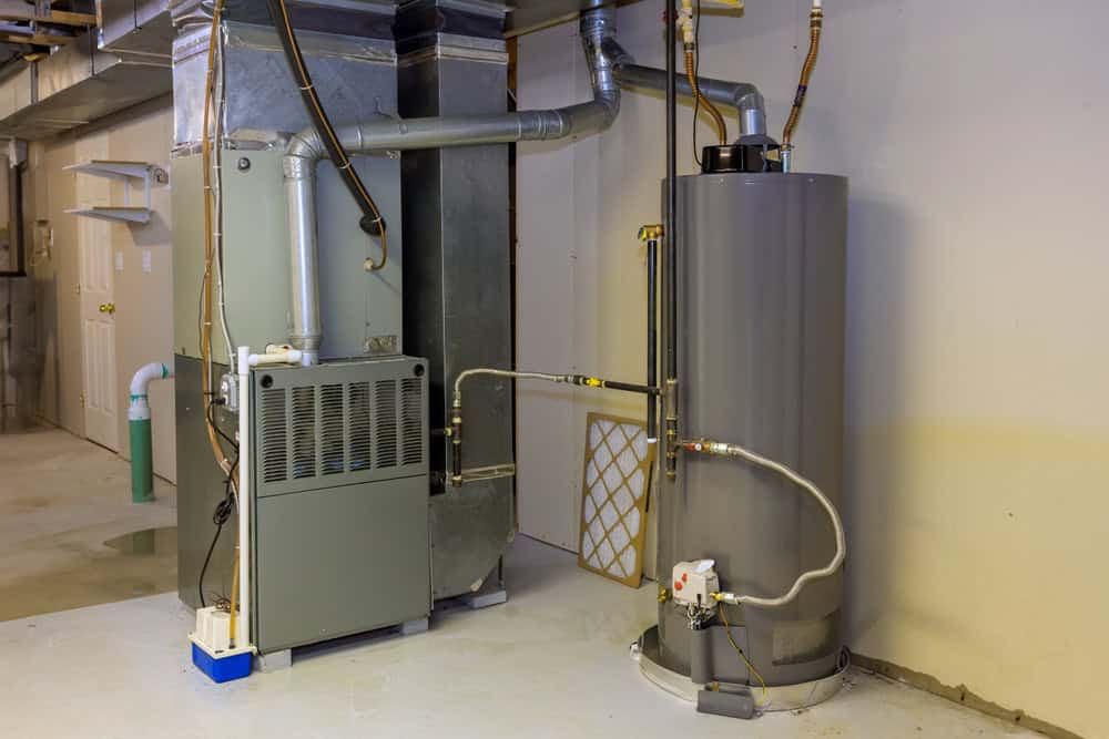 A residential utility room with a water heater and furnace, both essential for smooth HVAC services. The water heater is a tall, cylindrical, gray unit with pipes extending from the top and side. The furnace is a rectangular, gray unit connected to ductwork, with a filter propped against it.