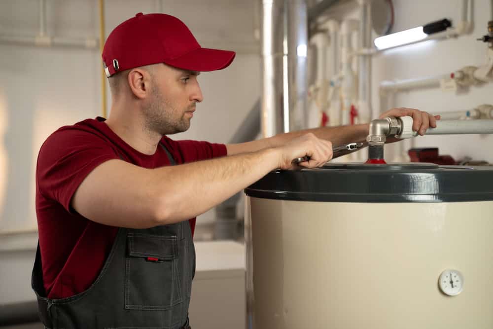 A man wearing a red cap and red shirt is diligently working on a water heater. Holding a wrench, he appears to be adjusting or repairing the unit. The background shows pipes and parts of the heating system, suggesting professional HVAC Services or potential Boiler Maintenance work.