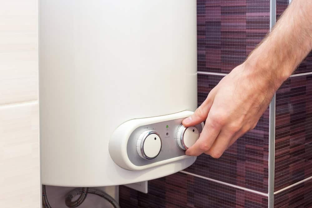 A person adjusts the settings on a white water heater, performing routine boiler maintenance on the unit mounted against a wall with brown tile. The water heater features two dials and a small display.