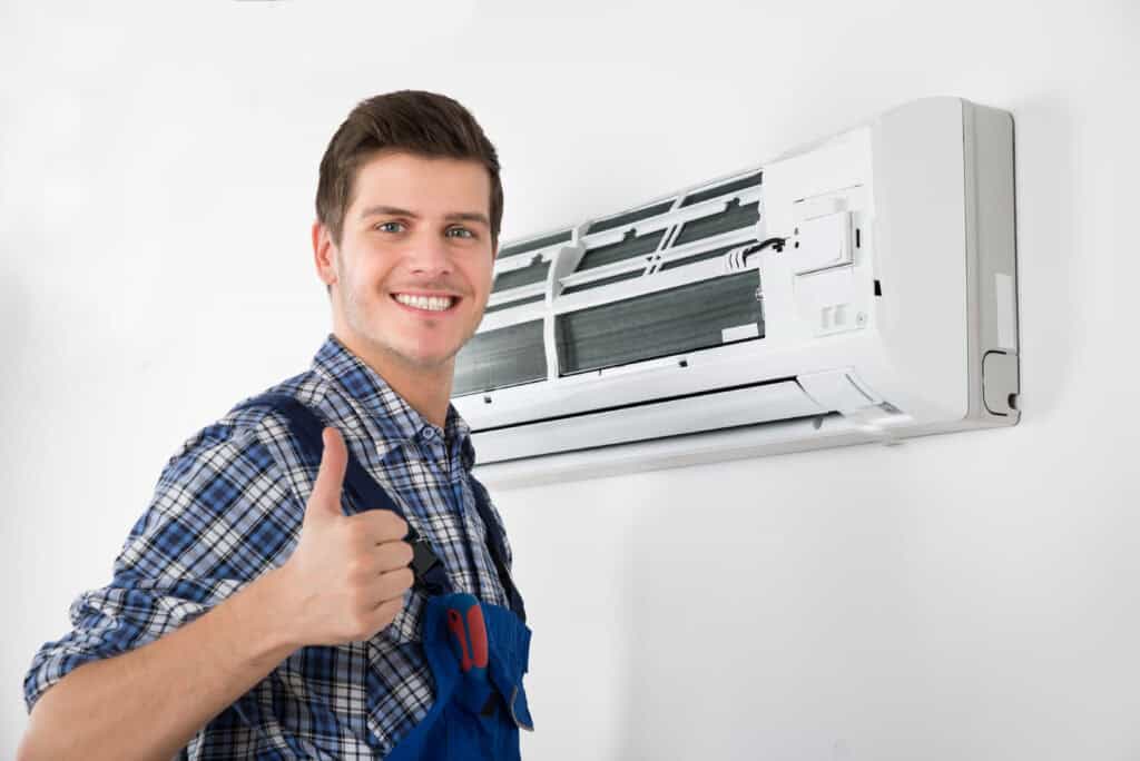 A person wearing a blue plaid shirt and blue overalls gives a thumbs-up in front of an open wall-mounted air conditioner. The person is smiling and appears to be happy with the AC repair done on the unit.