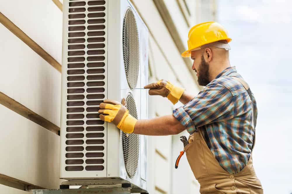 A technician wearing a yellow hard hat, gloves, and a plaid shirt is standing on a ladder while working on an outdoor air conditioning unit mounted on the side of a building. He is using a screwdriver to perform routine AC repair maintenance on the unit.